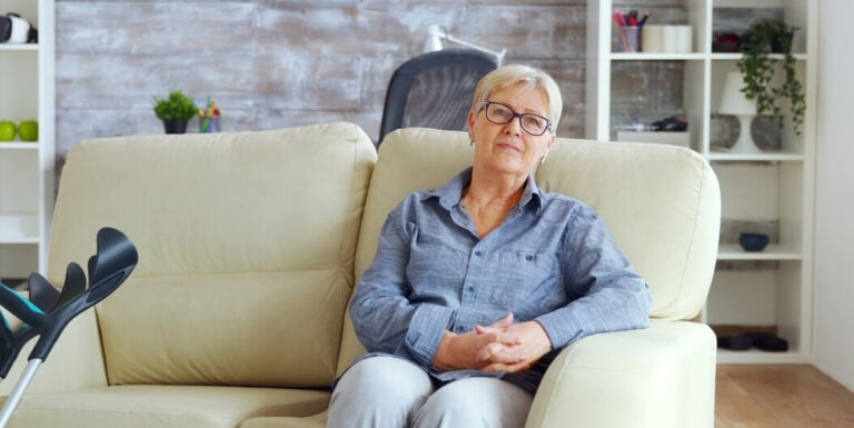 Elderly Woman Sitting on a Couch Beside a Crutch in a Calm Room at a Memory Care Facility